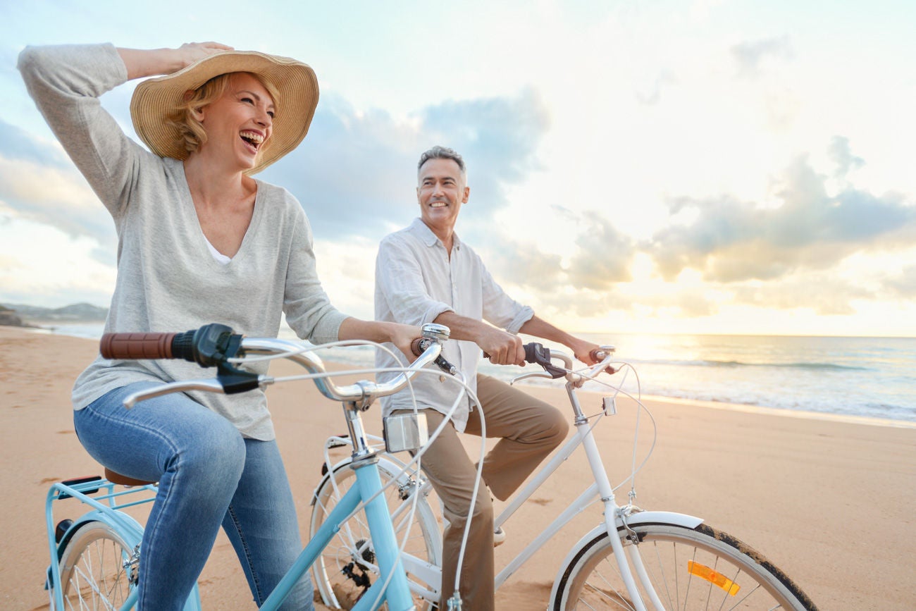 Mature couple cycling on the beach at sunset or sunrise. They are laughing and having fun. They are casually dressed. Could be a retirement vacation.